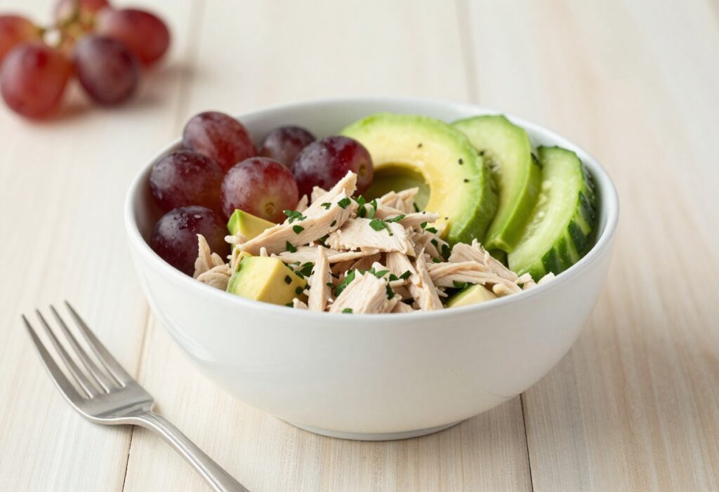 Bowl of canned chicken salad with avocado, cucumber, grapes, and herbs on a wooden table.