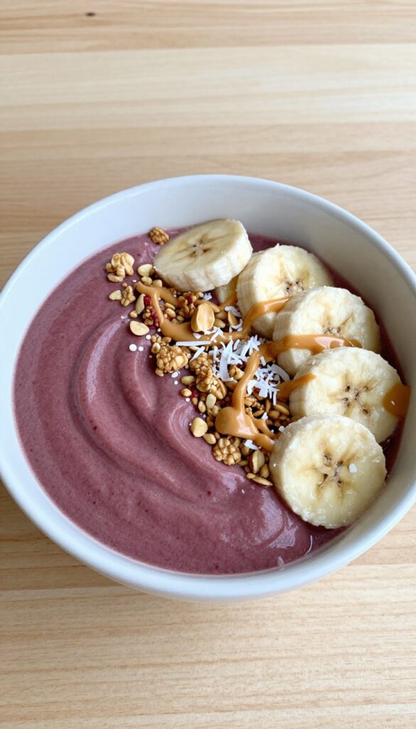Peanut butter and jelly smoothie bowl with granola, banana, coconut, and peanut butter drizzle on a wooden table.