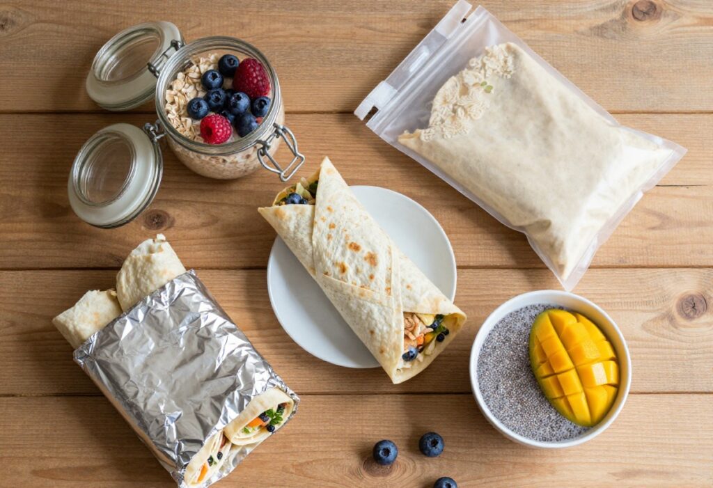 Meal-prepped breakfast items on a wooden table including overnight oats, burritos, smoothie pack, and chia pudding.